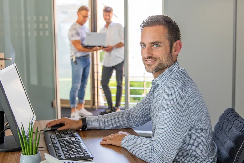 Cowirker arbeitet an einem Schreibtisch im Private Office, während zwei CoWirkerInnen im Hintergrund an einem Laptop stehen.