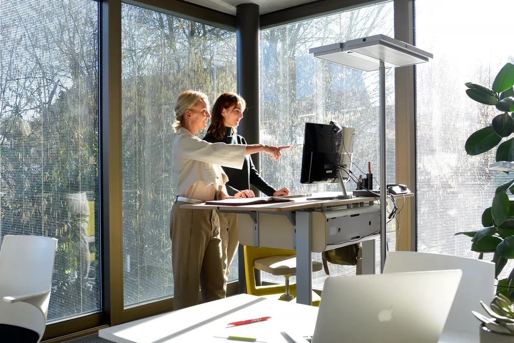 Zwei Frauen besprechen gemeinsam Projekte an einem höhenverstellbaren Schreibtisch im lichtdurchfluteten Coworking Space.